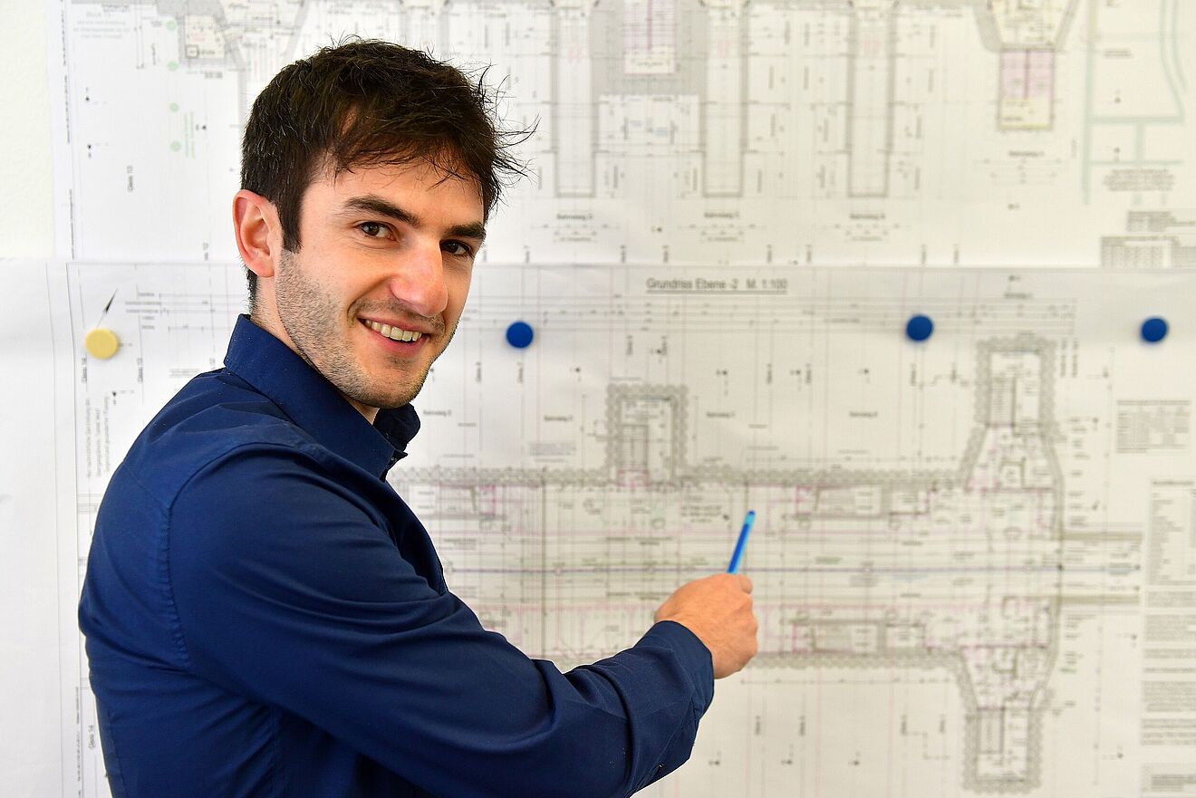 Mann zeigt mit einem Stift auf einen Papierplan an der Wand. Link zu einem Pop-Up mit Informationen zur Betreuung von Projekten.