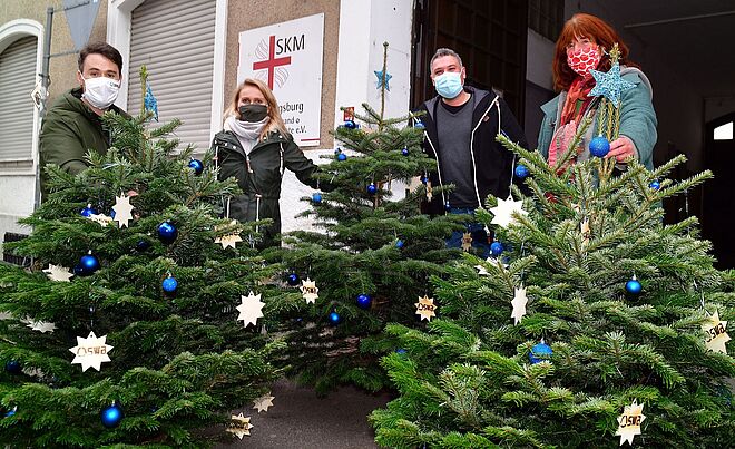 Vier Personen halten geschmückte Weihnachtsbäume vor dem SKM-Gebäude