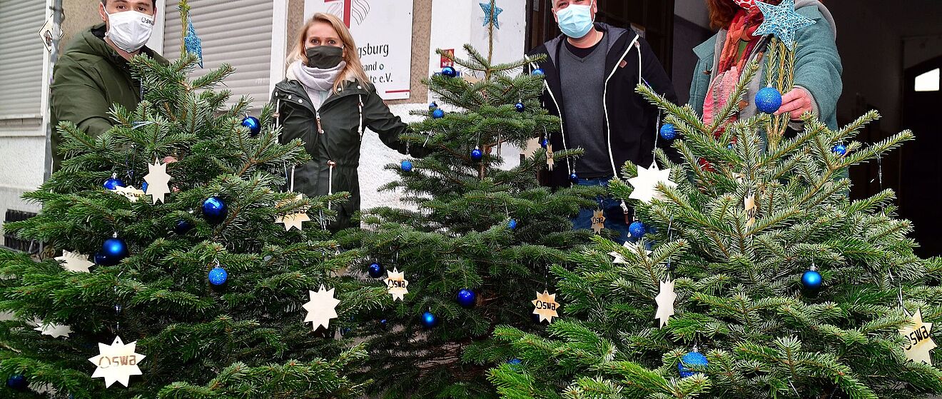Vier Personen halten geschmückte Weihnachtsbäume vor dem SKM-Gebäude
