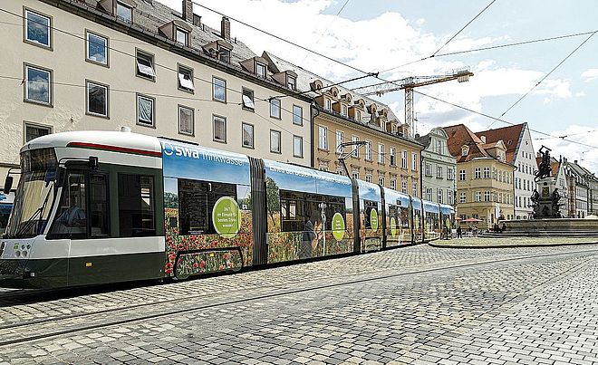Eine Straßenbahn der Stadtwerke Augsburg (swa) mit einer Blumen- und Wasserwerbung fährt auf Kopfsteinpflastergleisen am Rathausplatz in Augsburg vorbei.
