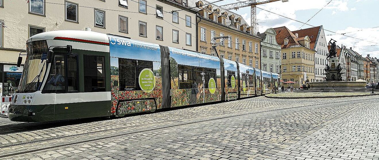 Eine Straßenbahn der Stadtwerke Augsburg (swa) mit einer Blumen- und Wasserwerbung fährt auf Kopfsteinpflastergleisen am Rathausplatz in Augsburg vorbei.
