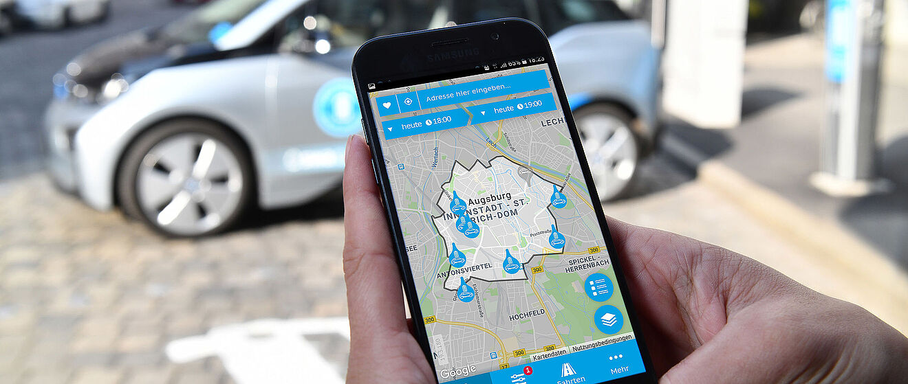Eine Hand hält ein Handy auf der die swa Mobil App geöffnet ist, im Hintergrund steht ein swa Carsharing Auto. - Ein Klick auf das Bild führt zur vergrößerten Fotoansicht.