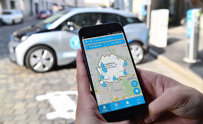 Eine Hand hält ein Handy auf der die swa Mobil App geöffnet ist, im Hintergrund steht ein swa Carsharing Auto. - Ein Klick auf das Bild führt zur vergrößerten Fotoansicht.