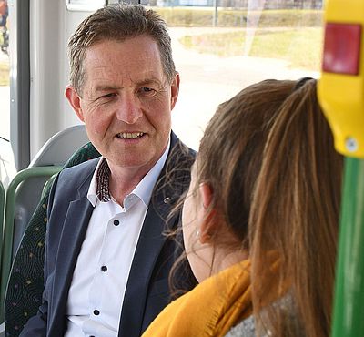 Ein Mann und eine Frau sitzen in der Straßenbahn und reden miteinander. Die Frau sitzt mit dem Rücken zur Kamera, der Mann hält einen Taschenrechner und Dokumente in der Hand.
