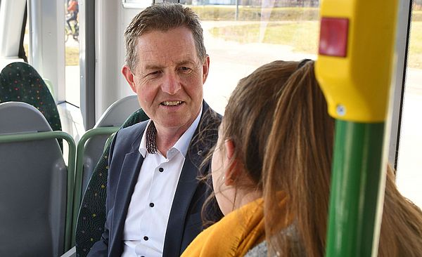 Ein Mann und eine Frau sitzen in der Straßenbahn und reden miteinander. Die Frau sitzt mit dem Rücken zur Kamera, der Mann hält einen Taschenrechner und Dokumente in der Hand.
