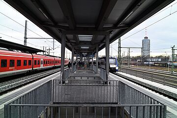Ein Bahnhofsgleis zeigt einen roten Regionalzug links und einen blau-weißen Zug rechts, die an einem Bahnsteig stehen. Eine offene, metallene Überführung führt nach unten, umgeben von Gleisen, Oberleitungen und einem hohen Gebäude im Hintergrund. Ein Klick auf das Bild führt zu einer größeren Ansicht.