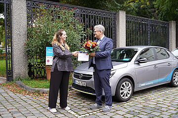 Ein Mann und eine Frau schütteln die Hände vor einem Carsharing Auto. Der Mann hält einen Blumenstrauß
