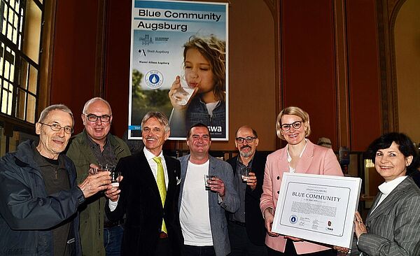 Eine Gruppe von Personen lächeln in die Kamera und halten Gläser mit Wasser in der Hand. Rechts stehen zwei Frauen, welche lächelnd ein Zertifikat halten.