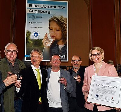 Eine Gruppe von Personen lächeln in die Kamera und halten Gläser mit Wasser in der Hand. Rechts stehen zwei Frauen, welche lächelnd ein Zertifikat halten.