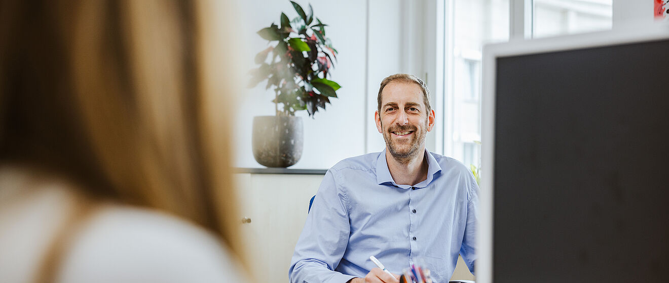 Mann in Hemd sitzt an einem Schreibtisch im Büro und grinst