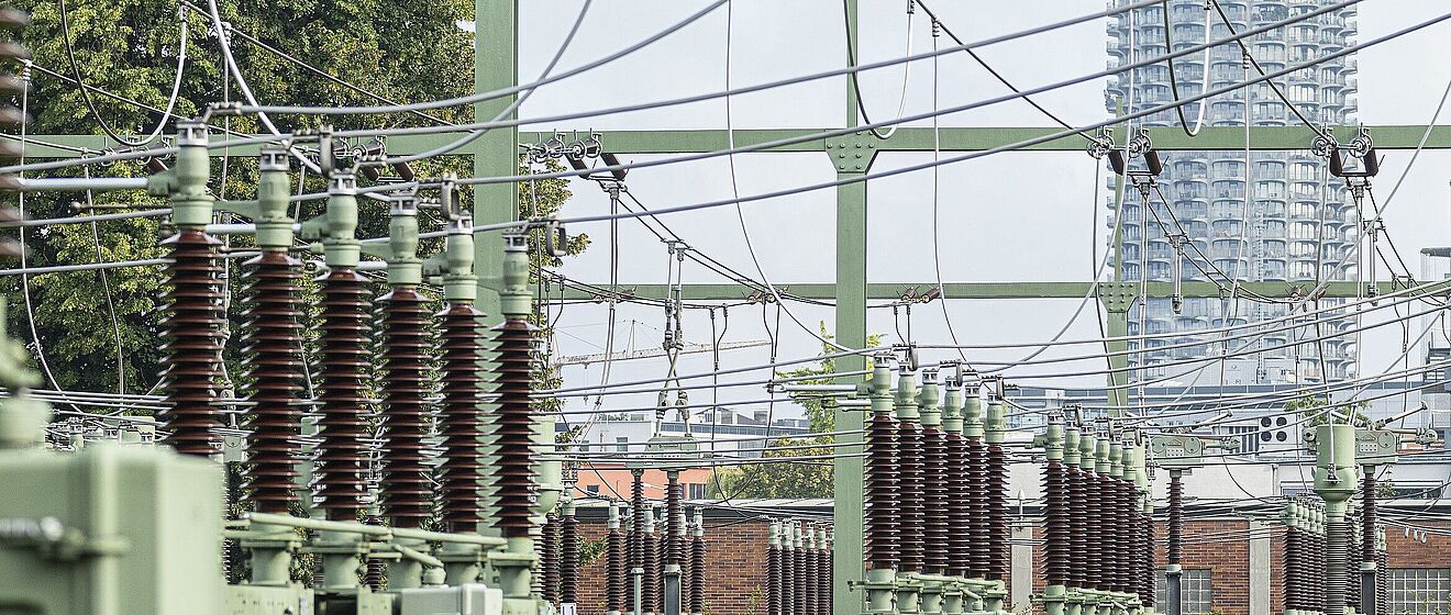 Im Vordergrund und mittig des Bildes sind die detailreichen, keramischen Isolatoren und die grünen Metallstrukturen eines Umspannwerks zu sehen, das Teil des Stromnetzes der Stadtwerke Augsburg (swa) ist. Im Hintergrund ragt das markante Hochhaus des Hotelturms Augsburg, auch bekannt als "Maiskolben", in den Himmel und bildet einen deutlichen urbanen Kontrast zur Industrieanlage.