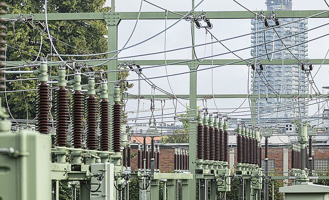 Im Vordergrund und mittig des Bildes sind die detailreichen, keramischen Isolatoren und die grünen Metallstrukturen eines Umspannwerks zu sehen, das Teil des Stromnetzes der Stadtwerke Augsburg (swa) ist. Im Hintergrund ragt das markante Hochhaus des Hotelturms Augsburg, auch bekannt als "Maiskolben", in den Himmel und bildet einen deutlichen urbanen Kontrast zur Industrieanlage.