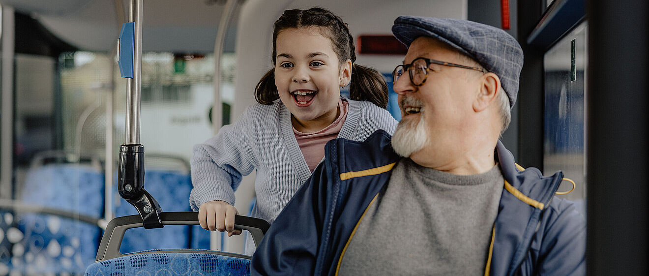 Ein älterer Mann sitzt in einem Bus und schaut nach hinten zu einem kleinen lachenden Mädchen
