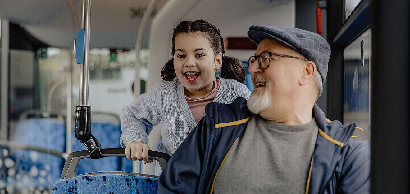 Ein älterer Mann sitzt in einem Bus und schaut nach hinten zu einem kleinen lachenden Mädchen