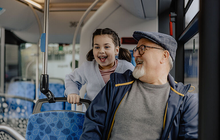 Ein älterer Mann sitzt in einem Bus und schaut nach hinten zu einem kleinen lachenden Mädchen