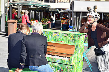 Zwei Frauen spielen zusammen auf dem bemalte Klavier und eine weitere Frau hört zu. - Beim anklicken wird das Bild vergrößert.