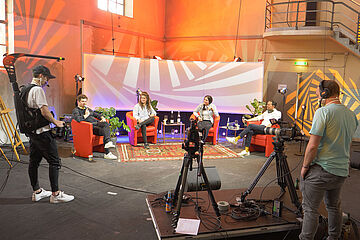 Vier Personen sitzen mit Mikrofonen auf einem roten Sessel vor ihnen steht eine Kameracrew mit mehreren Kameras. - Ein Klick auf das Bild führt zur vergrößerten Fotoansicht.