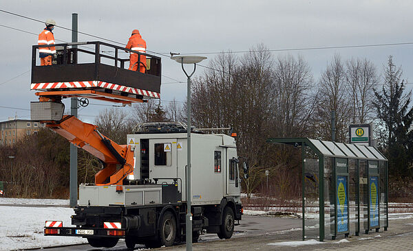 Zwei Männer in orangener Schutzkleidung stehen auf einem Personenaufzug und arbeiten an einer Stromoberleitung. Im Hintergrund ist eine Haltestelle.