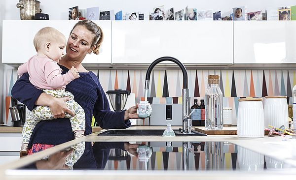 Frau steht mit ihrem Baby in der Küche und füllt Wasser in seine Flasche.