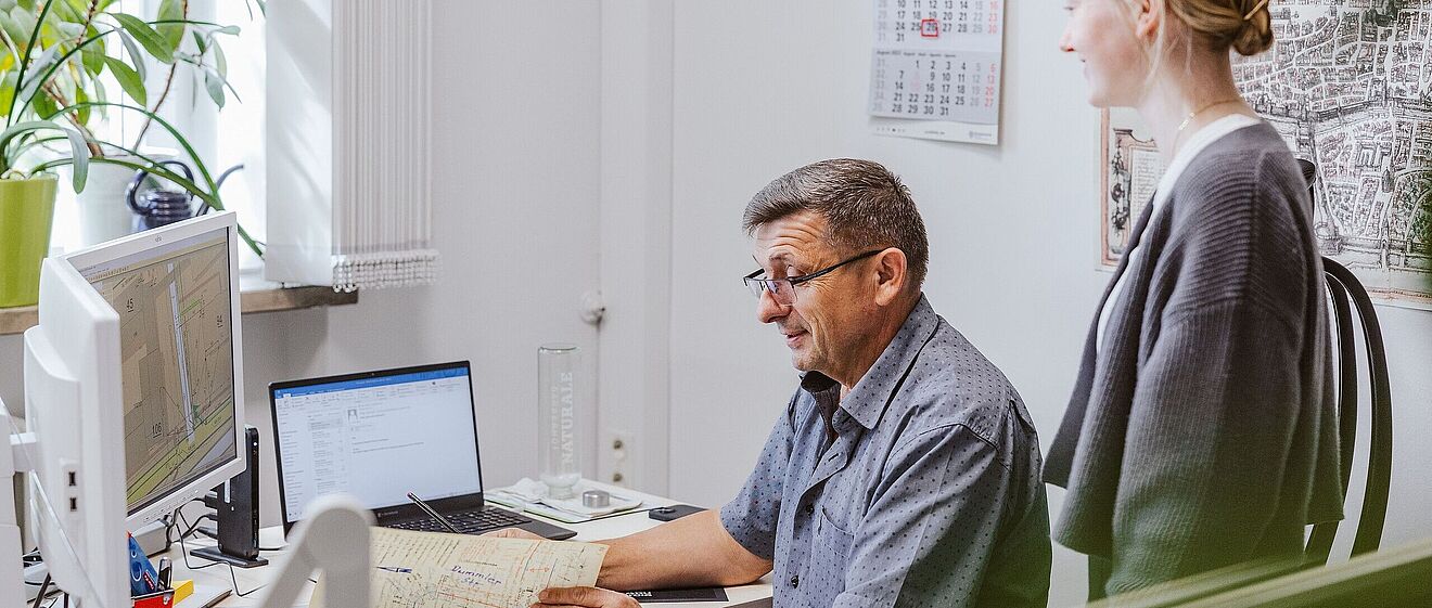 Mann sitzt am Schreibtisch vor Monitoren und einem Laptop und betrachtet Notizen während eine Frau hinter ihm steht