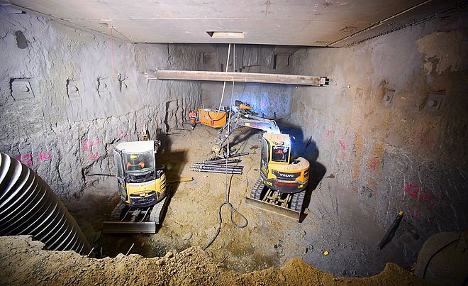 Eine unterirdische Baustelle zeigt zwei kleine, gelbe Bagger und weitere orangefarbene Maschinen. Der beengte Bereich umfasst rohe Betonwände und Erdreich.
