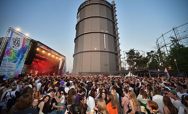 Das Bild zeigt eine feiernde Menschenmenge vor einer großen Bühne. Die Bühne ist mit Scheinwerfern rot beleuchtet. Links neben der Bühne ist ein Banner zu sehen, auf dem Modular Festival steht. Auf dem Banner kann man lesen, dass das Festival von Sponsoren wie den Stadtwerken Augsburg (swa) gefördert wird. Im Hintergrund des Bilds ist der Gaskessel zu sehen.
