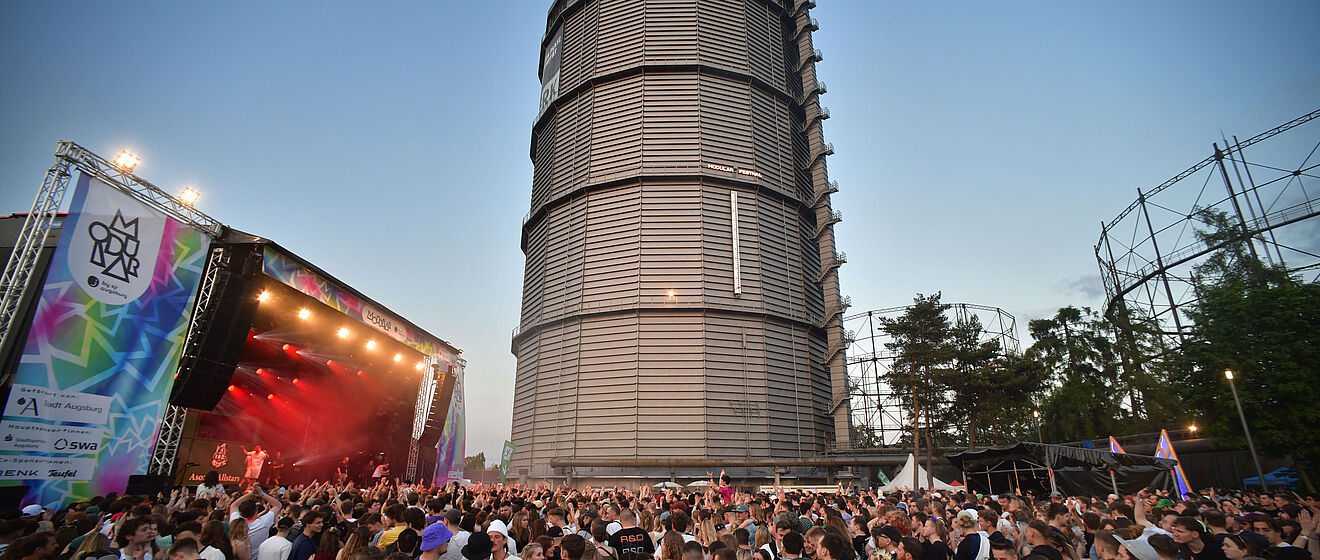 Das Bild zeigt eine feiernde Menschenmenge vor einer großen Bühne. Die Bühne ist mit Scheinwerfern rot beleuchtet. Links neben der Bühne ist ein Banner zu sehen, auf dem Modular Festival steht. Auf dem Banner kann man lesen, dass das Festival von Sponsoren wie den Stadtwerken Augsburg (swa) gefördert wird. Im Hintergrund des Bilds ist der Gaskessel zu sehen.