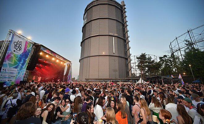Das Bild zeigt eine feiernde Menschenmenge vor einer großen Bühne. Die Bühne ist mit Scheinwerfern rot beleuchtet. Links neben der Bühne ist ein Banner zu sehen, auf dem Modular Festival steht. Auf dem Banner kann man lesen, dass das Festival von Sponsoren wie den Stadtwerken Augsburg (swa) gefördert wird. Im Hintergrund des Bilds ist der Gaskessel zu sehen.