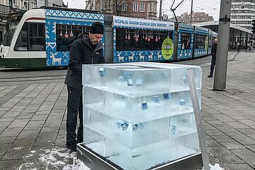 Ein Mann in schwarzer Kleidung bearbeitet mit seinen Händen einen mehrstufigen, glasklaren Eisblock, in den blaue Objekte eingefroren sind. Der Block steht auf einem gepflasterten Stadtplatz, während im Hintergrund eine weihnachtlich geschmückte Straßenbahn der Stadtwerke Augsburg mit der Aufschrift 