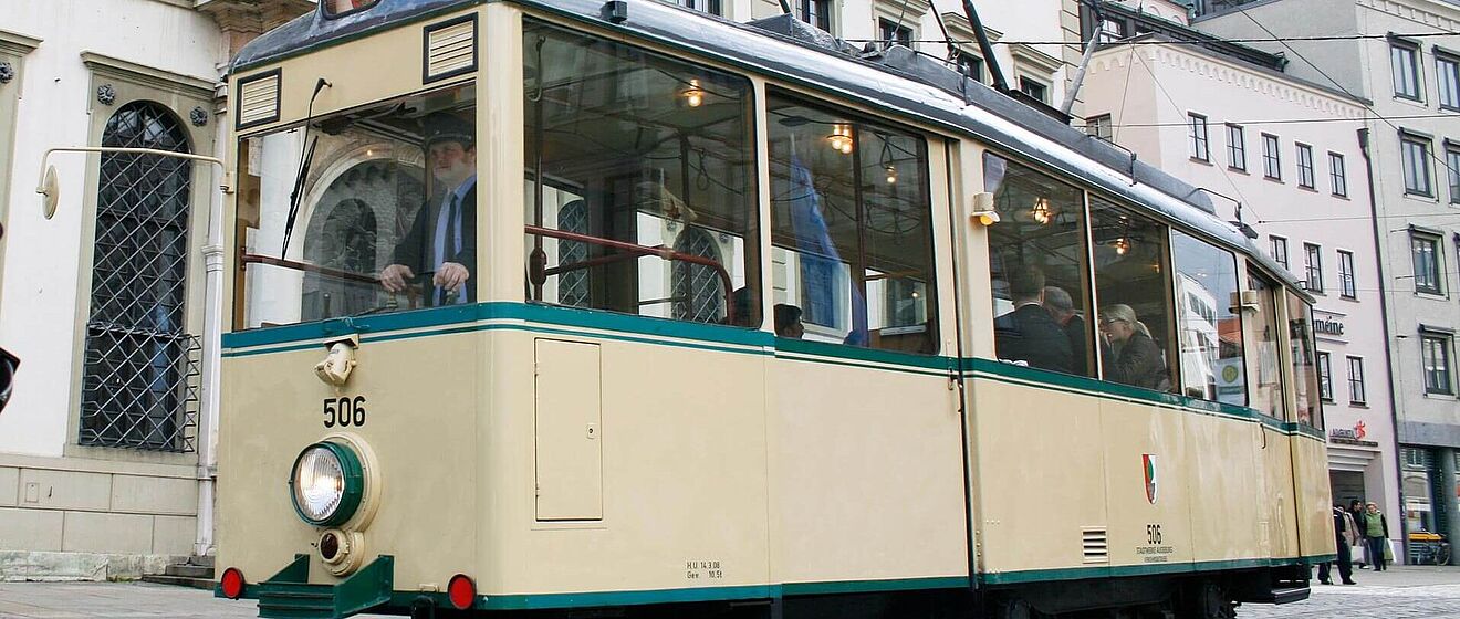 Eine historische Straßenbahn mit einem Fahrer und sitzenden Passagieren 