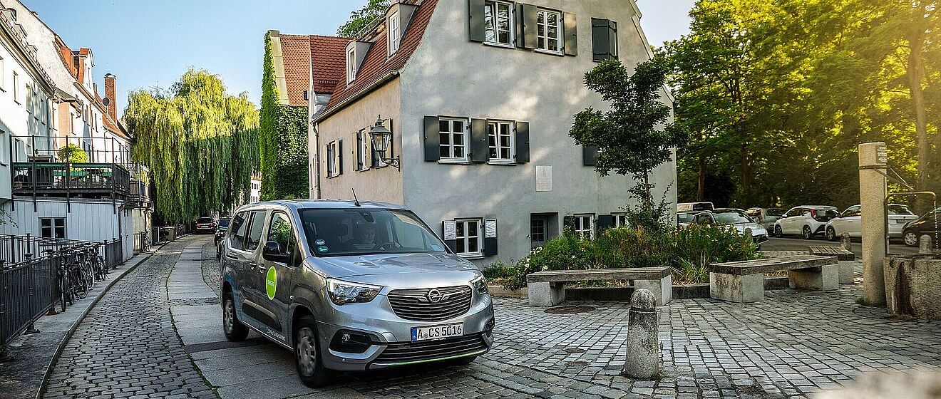 Fahrer sitzt im swaxi in der Augsburger Altstadt. Rechts ist ein swa Trinkwasserbrunnen. Im Hintergrund sind Gebäude.