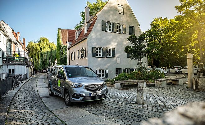 Fahrer sitzt im swaxi in der Augsburger Altstadt. Rechts ist ein swa Trinkwasserbrunnen. Im Hintergrund sind Gebäude.