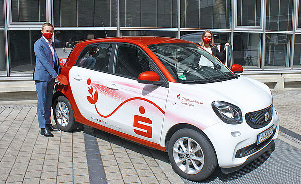 Ein Auto der Marke Smart, in dass eine Frau mit Mundschutz einsteigt und einem Herrn, der das Auto zum Laden anschließt. Auf dem Auto ist das Stadtsparkassen Symbol. 
