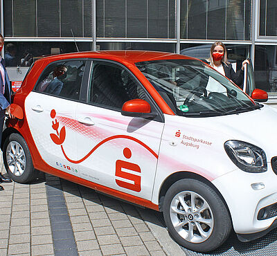 Ein Auto der Marke Smart, in dass eine Frau mit Mundschutz einsteigt und einem Herrn, der das Auto zum Laden anschließt. Auf dem Auto ist das Stadtsparkassen Symbol. 