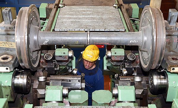 Ein Arbeiter im Blaumann und gelbem Schutzhelm hockt unter einer großen, grünen Radsatzdrehmaschine und führt Wartungsarbeiten an den massiven, grauen Eisenbahnrädern durch.