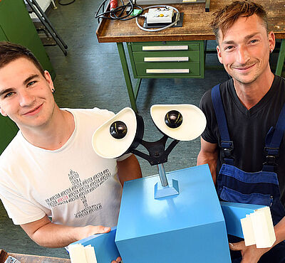 Zwei Männer stehen nebeneinander an einer Werkbank und halten Swally in die Kamera.