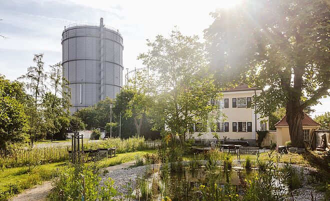 Gaskessel im Gaswerk. Im Vordergrund sind Bäume und ein Gebäude.