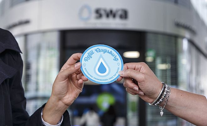 Vor dem swa Kundencenter am Königsplatz wird ein Aufkleber mit der Aufschrift Refill Augsburg von zwei Händen gehalten