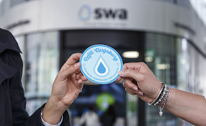 Vor dem swa Kundencenter am Königsplatz wird ein Aufkleber mit der Aufschrift Refill Augsburg von zwei Händen gehalten