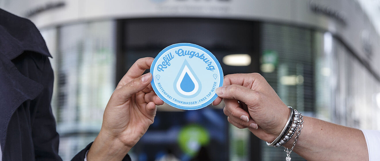 Vor dem swa Kundencenter am Königsplatz wird ein Aufkleber mit der Aufschrift Refill Augsburg von zwei Händen gehalten
