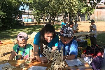 Zwei Kinder und eine erwachsene Person basteln im Freien an einem Tisch Holz-Schmetterlinge. Im Hintergrund ist das Gaswerk Augsburg zu sehen. Ein Klick auf das Bild führt zu einer größeren Ansicht.