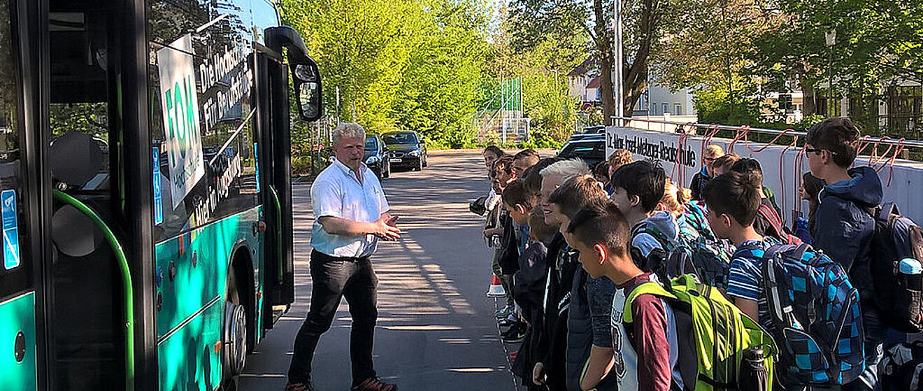 Auf der rechten Seite befindet sich eine Schulklasse sowie eine Mauer der Dr.-Max-Josef-Metzger-Realschule. Auf der linken Seite steht ein Mann vor einem Bus.