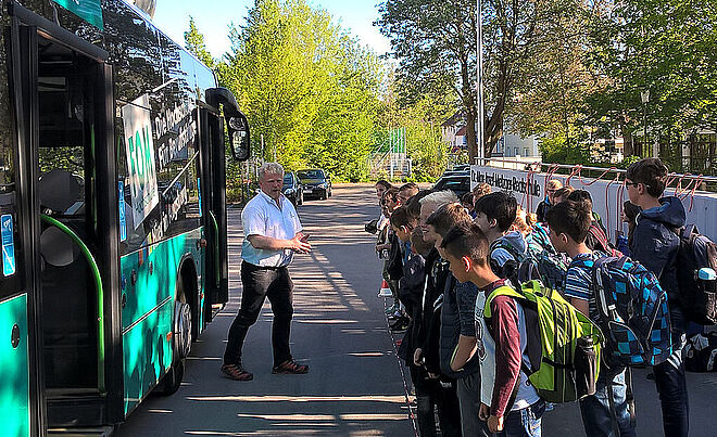 Auf der rechten Seite befindet sich eine Schulklasse sowie eine Mauer der Dr.-Max-Josef-Metzger-Realschule. Auf der linken Seite steht ein Mann vor einem Bus.