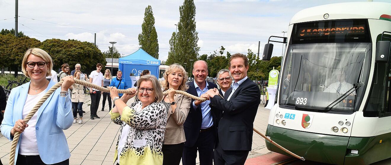 Im Vordergrund ziehen mehrere Personen symbolisch an einem dicken Seil, das mit einer grünen und weißen Straßenbahn des Typs Bombardier Flexity Outlook C der Stadtwerke Augsburg verbunden ist, welche die Wagennummer 893 trägt.