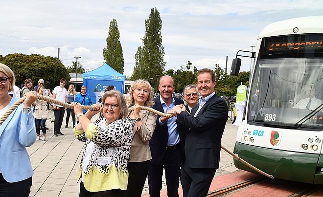 Im Vordergrund ziehen mehrere Personen symbolisch an einem dicken Seil, das mit einer grünen und weißen Straßenbahn des Typs Bombardier Flexity Outlook C der Stadtwerke Augsburg verbunden ist, welche die Wagennummer 893 trägt.