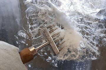 Ein mit Arbeitshandschuhen ausgestatteter Arm verwendet einen Eispickel, um in einen großen Eisblock zu hacken. Ein Klick auf das Bild führt zu einer größeren Ansicht.