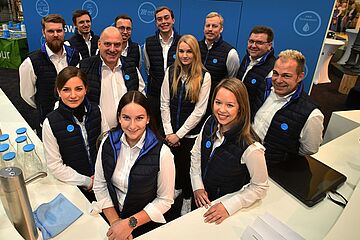 Eine Gruppe von 12 Personen, die meisten tragen dunkle Westen mit blauen Akzenten, steht hinter einem weißen Messetresen und posiert für ein Foto. Ein Klick auf das Bild führt zu einer größeren Ansicht.