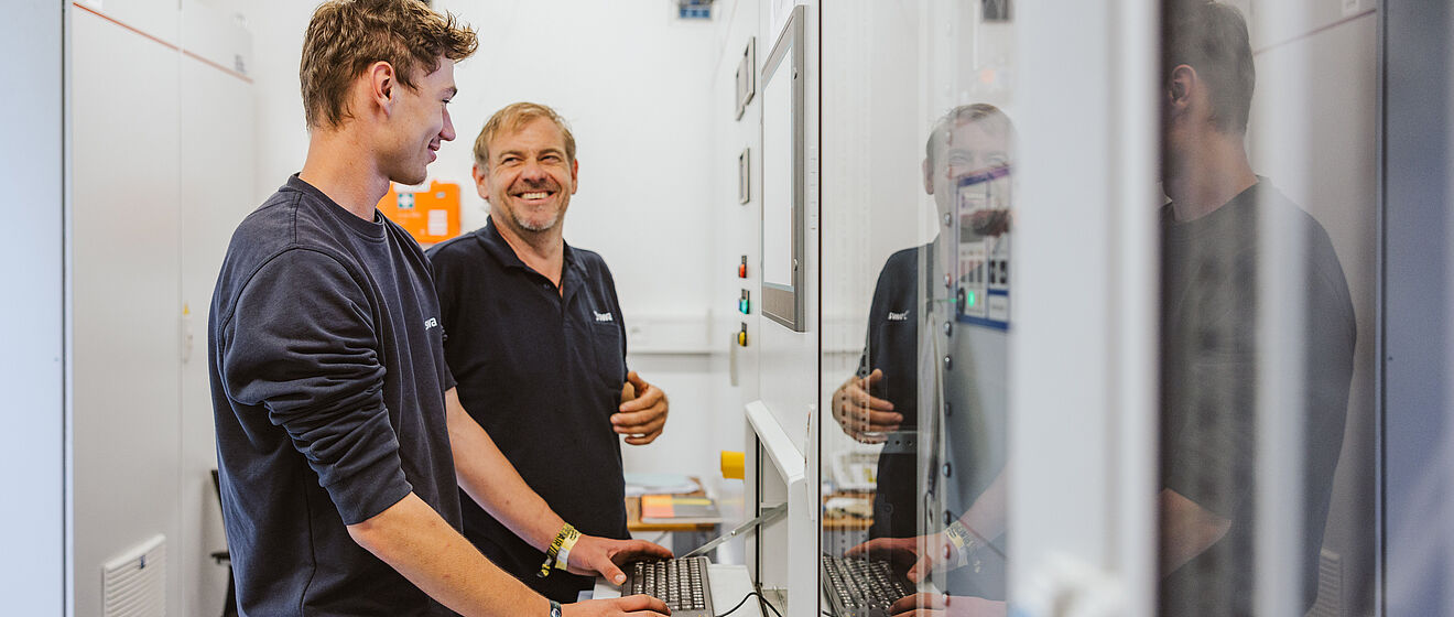 Zwei lachende Elektriker mit swa Pullover im Keller vor einem Bildschirm und Schaltern