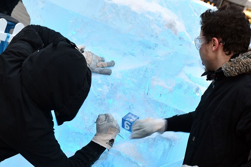 Zwei Männer, bekleidet mit schwarzen Jacken, Arbeitshandschuhen und Schutzbrillen, hacken mit ihren Händen und möglicherweise einem Eispickel auf einen großen, beleuchteten Eisblock ein. Ein kleiner blauer Würfel mit der Nummer "6" ist prominent im Eis eingefroren.