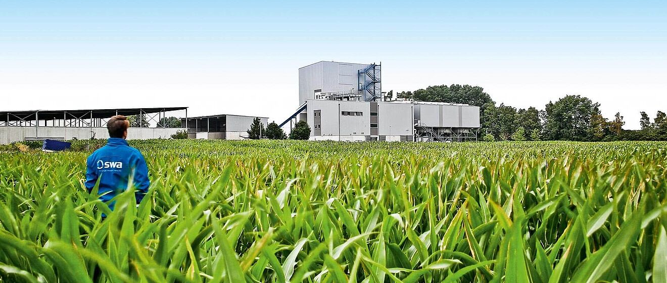 Mann mit blauer swa Jacke steht in einem Feld und schaut zum Biomasse Heizkraftwerk im Hintergrund
